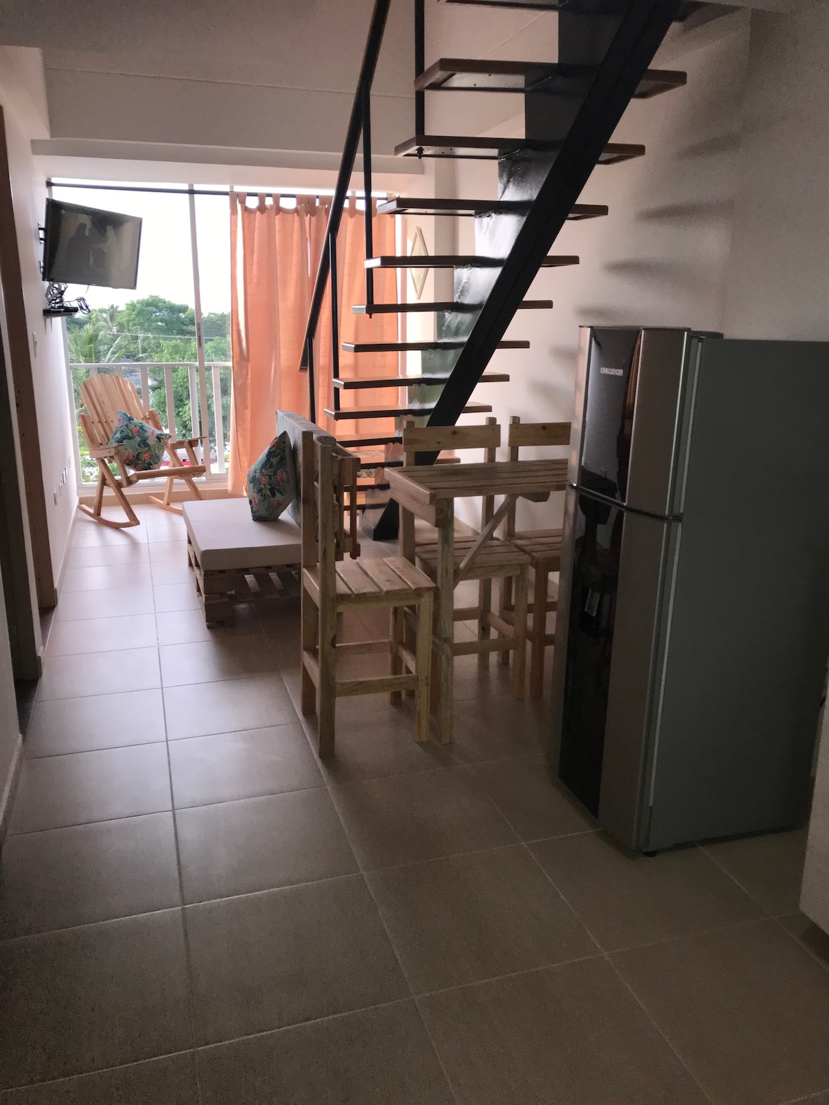 A light-filled space showcases a wooden dining set, accompanied by a refrigerator. A staircase with a sleek black railing leads to the upper level. Comfortable chairs are positioned near large windows, with soft curtains allowing natural light to illuminate the area.