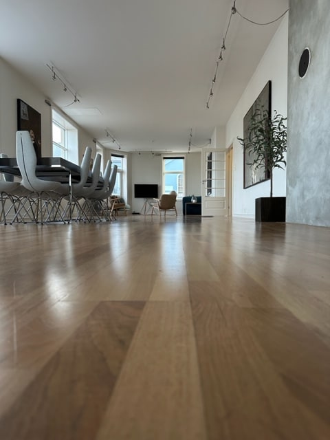 A spacious interior is featured, showcasing a light wood floor that extends towards a dining area with gray chairs. Large windows allow natural light to fill the room, highlighting a minimalist design that includes a modern television and artwork on the walls.