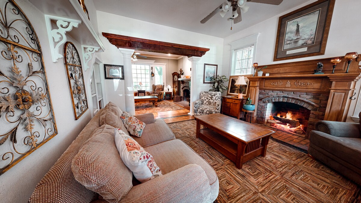 A cozy living area features a light-colored sectional sofa, complemented by a wooden coffee table. A warm fire is glowing in a brick fireplace, framed by artwork and decorative accents. Natural light enters through multiple windows, enhancing the inviting ambiance.