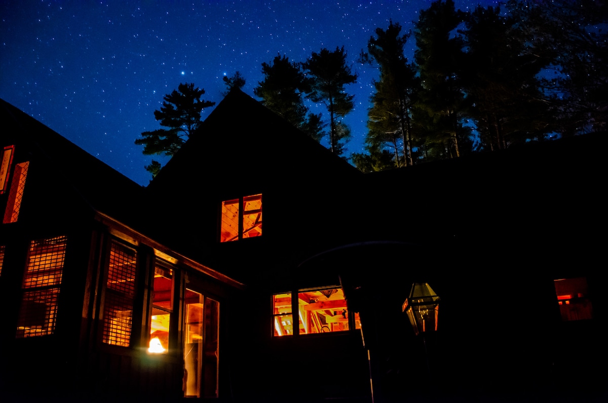 A cozy lodge is illuminated against a backdrop of a starry night sky. The structure showcases warm light shining through large windows, hinting at a welcoming interior. Dark silhouettes of trees frame the scene, enhancing the serene atmosphere of the natural surroundings.