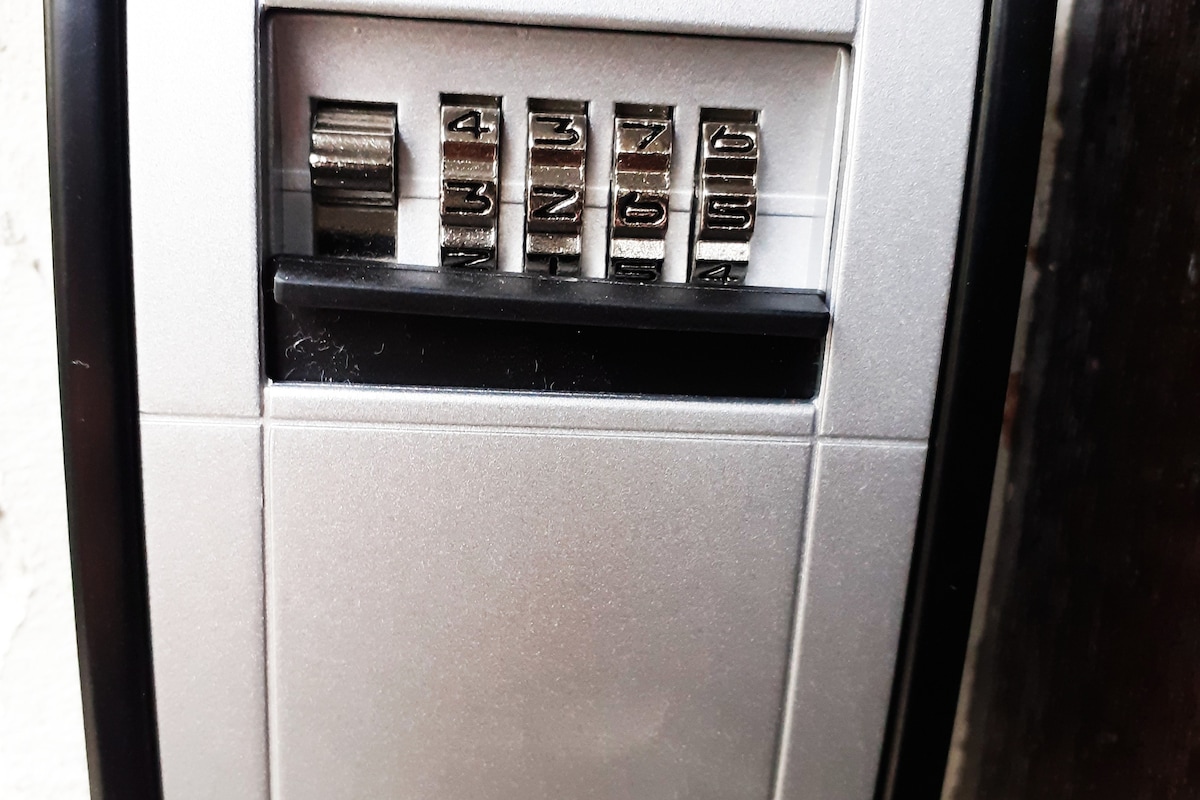 A lockbox is shown, featuring a series of numbered dials. Each dial is clearly visible, with its numbers displayed for code entry. The box is mounted against a contrasting wall, providing a practical access point for guests.