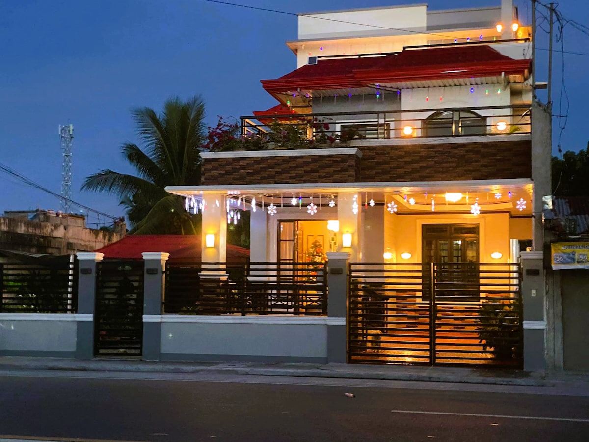 A modern two-story house is depicted, showcasing a welcoming facade adorned with decorative lights. The structure features a red roof, balcony with greenery, and horizontal black slatted fencing. The exterior is illuminated with soft yellow lighting, creating a warm evening ambiance.