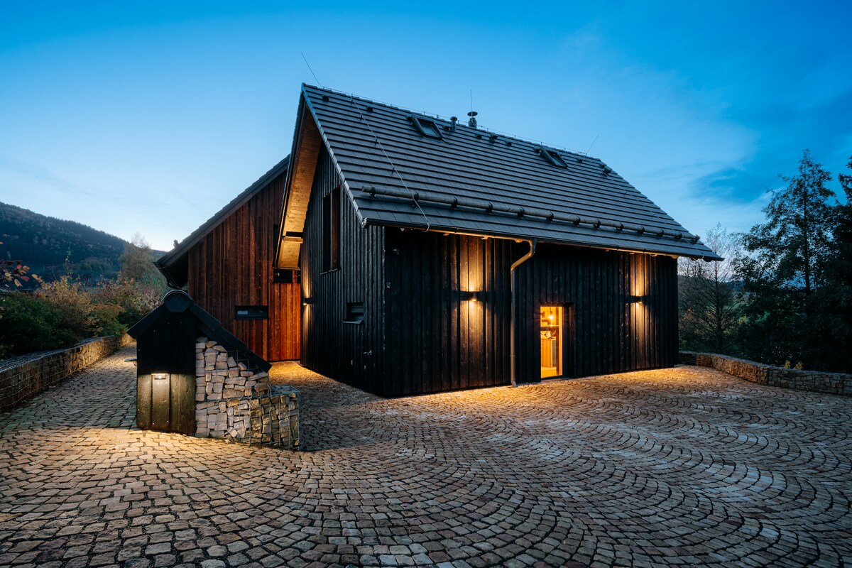The exterior of a modern, wooden chalet is presented, featuring a steeply pitched roof and large windows. Warm lighting from wall fixtures illuminates the dark wooden façade, while a circular stone pathway leads up to the entrance. Trees and hills create a natural backdrop.