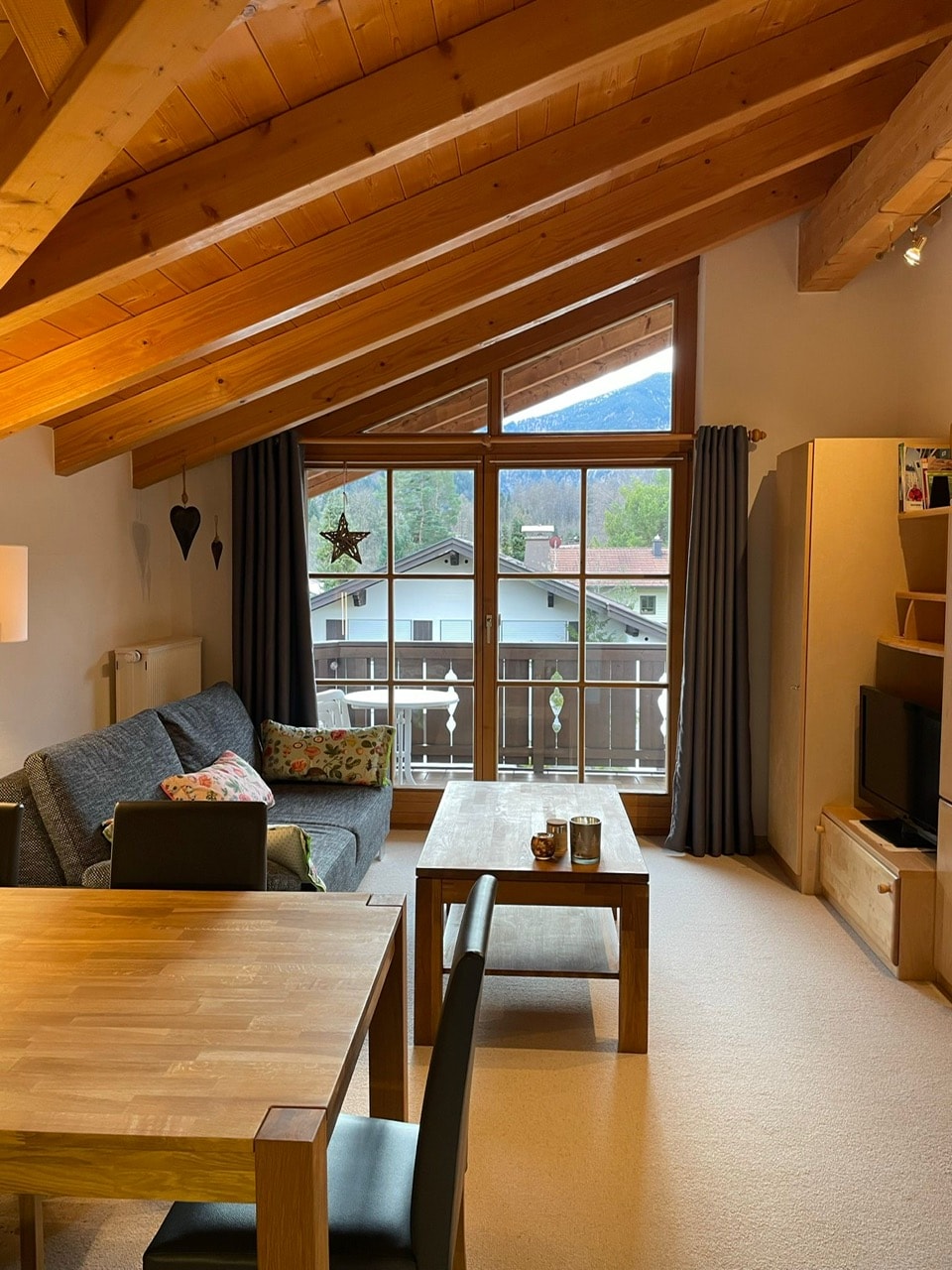 A bright living area features a sloped wooden ceiling and large windows, allowing for natural light. Comfortable seating arrangements include a gray sofa and two chairs around a wooden dining table. The decor includes soft cushions and a small television unit, creating a cozy gathering space.