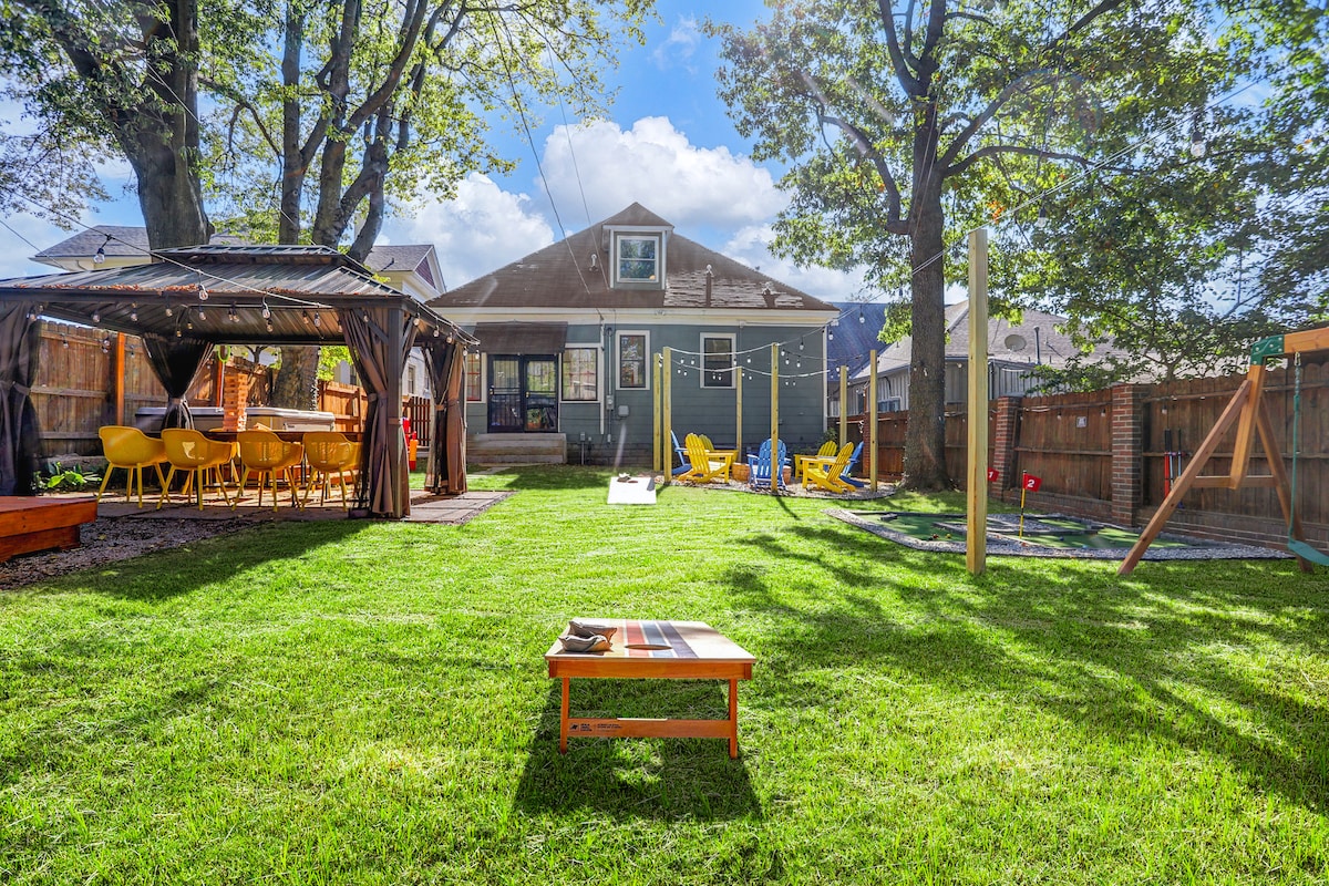 A spacious backyard is highlighted with bright green grass and scattered sunny seating areas. Yellow lounge chairs and a dining area with string lights are visible. A wooden table is placed in the foreground. A play structure and a covered patio are noticeable in the background.