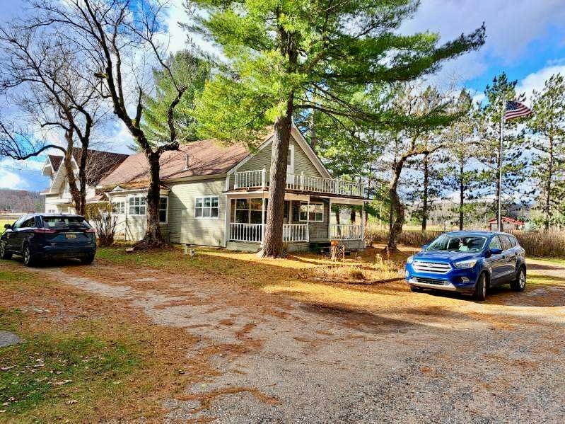 The exterior of the home is visible, featuring a smooth gravel driveway leading towards the entrance. Two parked cars are positioned beside the house, with tall trees providing shade. A porch is seen extending from the house, and a flag gently flutters nearby.