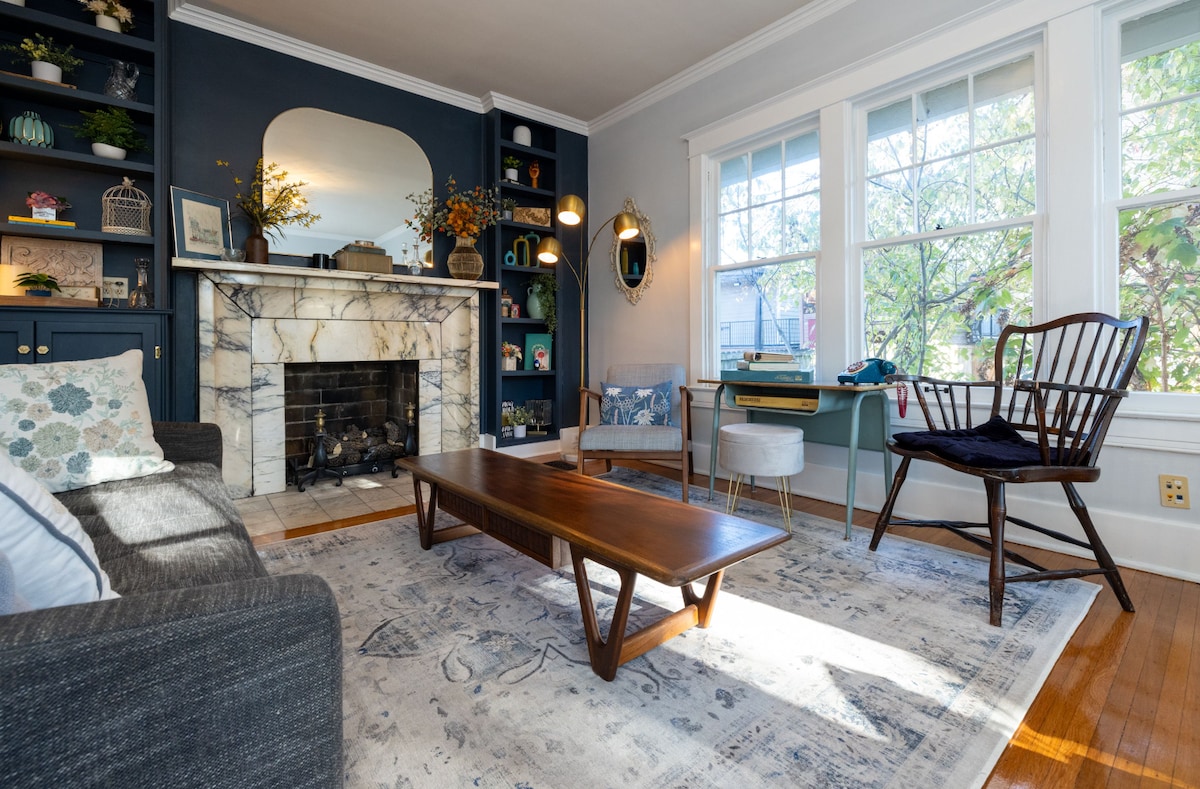 A stylish living room features a marble fireplace with decorative items above it. A wooden coffee table sits on a soft area rug, while a dark gray sofa and a classic wooden chair provide seating. Large windows offer natural light and views of greenery outside.
