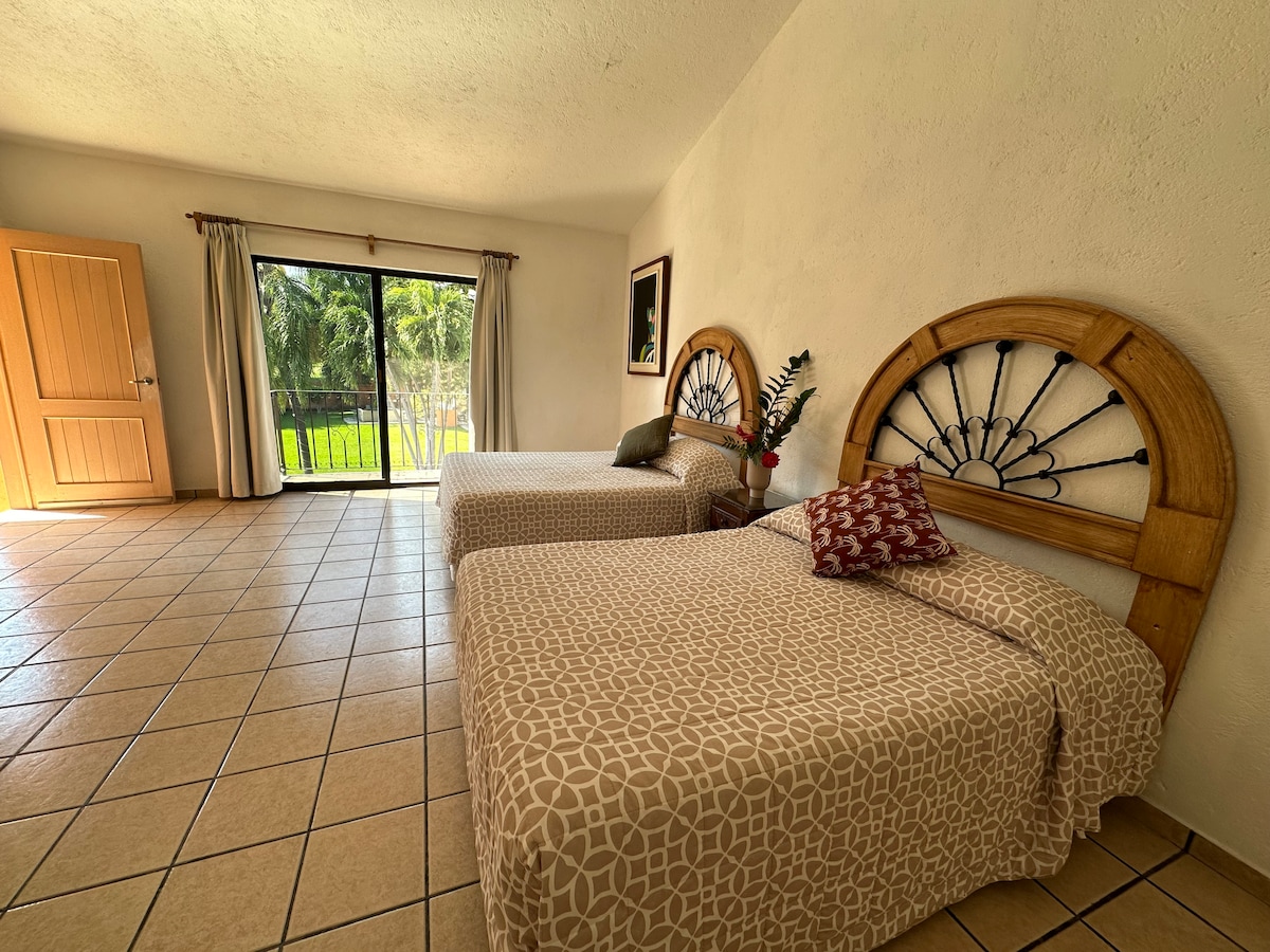 The spacious room features two twin beds with intricate wooden headboards, each dressed in patterned beige bedding. A large glass door allows natural light to fill the space, offering views of the green outdoor area. A decorative plant and a small throw pillow add a touch of warmth.