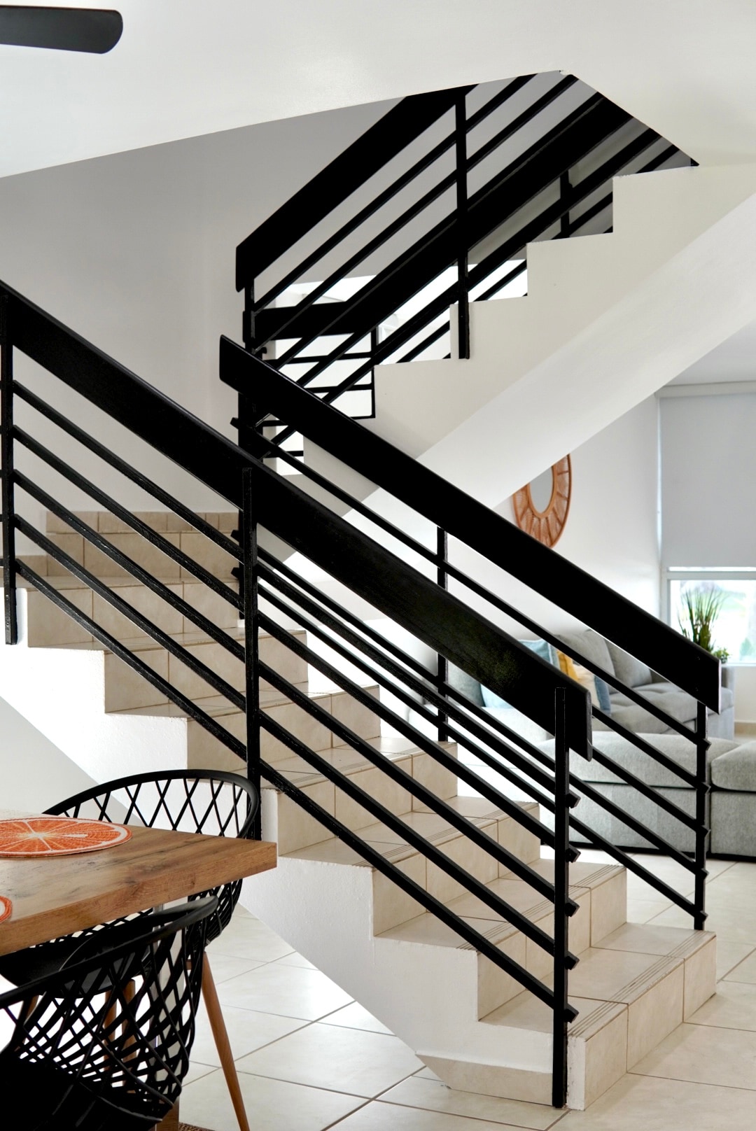 A modern staircase with sleek black railings is prominently featured. The light-colored tiles contrast with the dark handrails, guiding the eye up to the upper level. A dining area with a wooden table is visible in the foreground, adding warmth to the open layout.