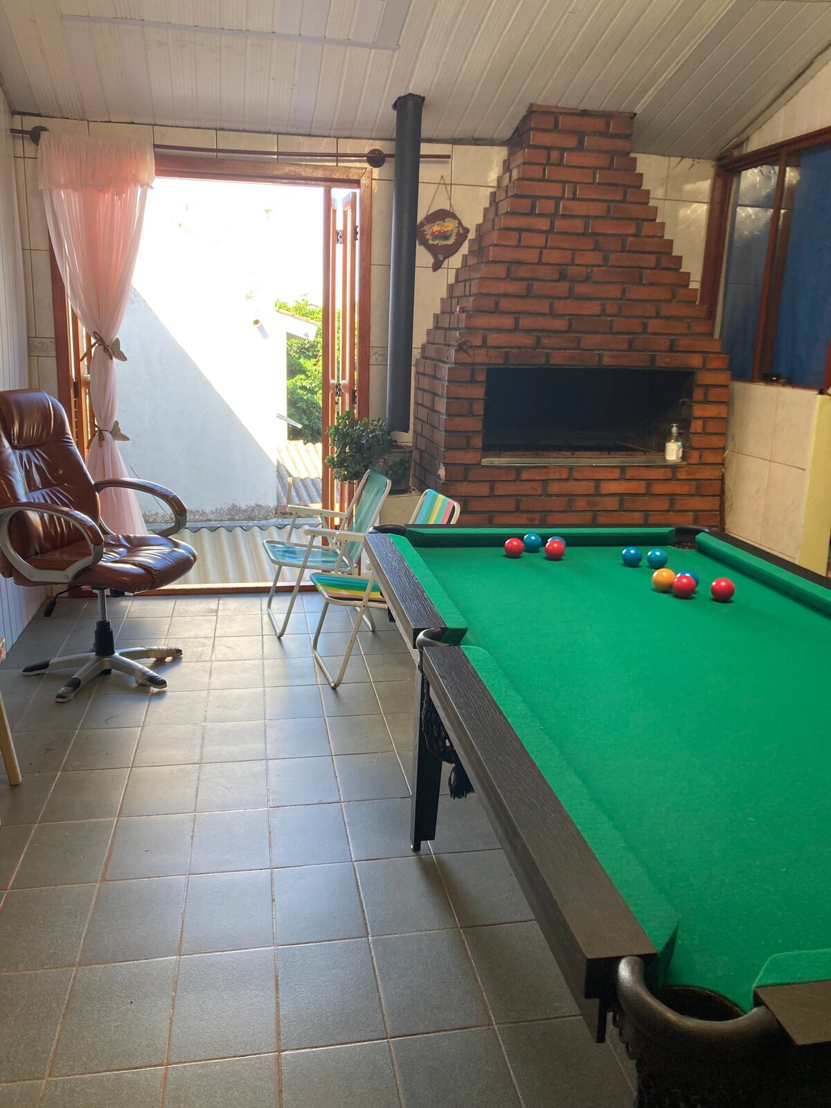 An inviting game room is featured, highlighted by a green billiard table with colorful balls arranged for play. A brick barbecue area stands nearby, with ample natural light entering through open double doors. Two chairs and a comfortable office chair are also visible.