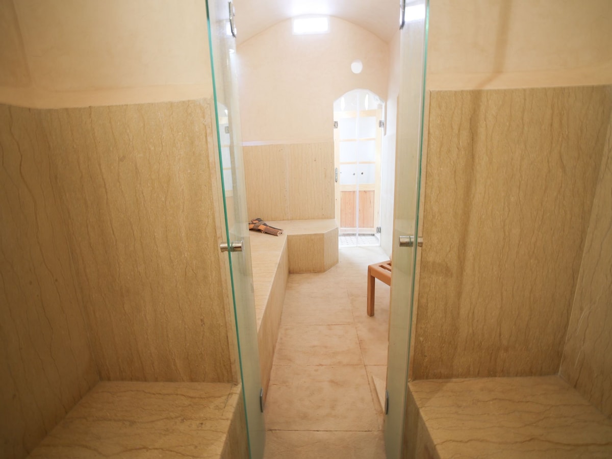 A spacious steam room features smooth stone walls and benches on either side. Light enters through a small window above, creating a serene atmosphere. The stone flooring complements the natural hues of the walls, and a wooden bench stands at the far end.