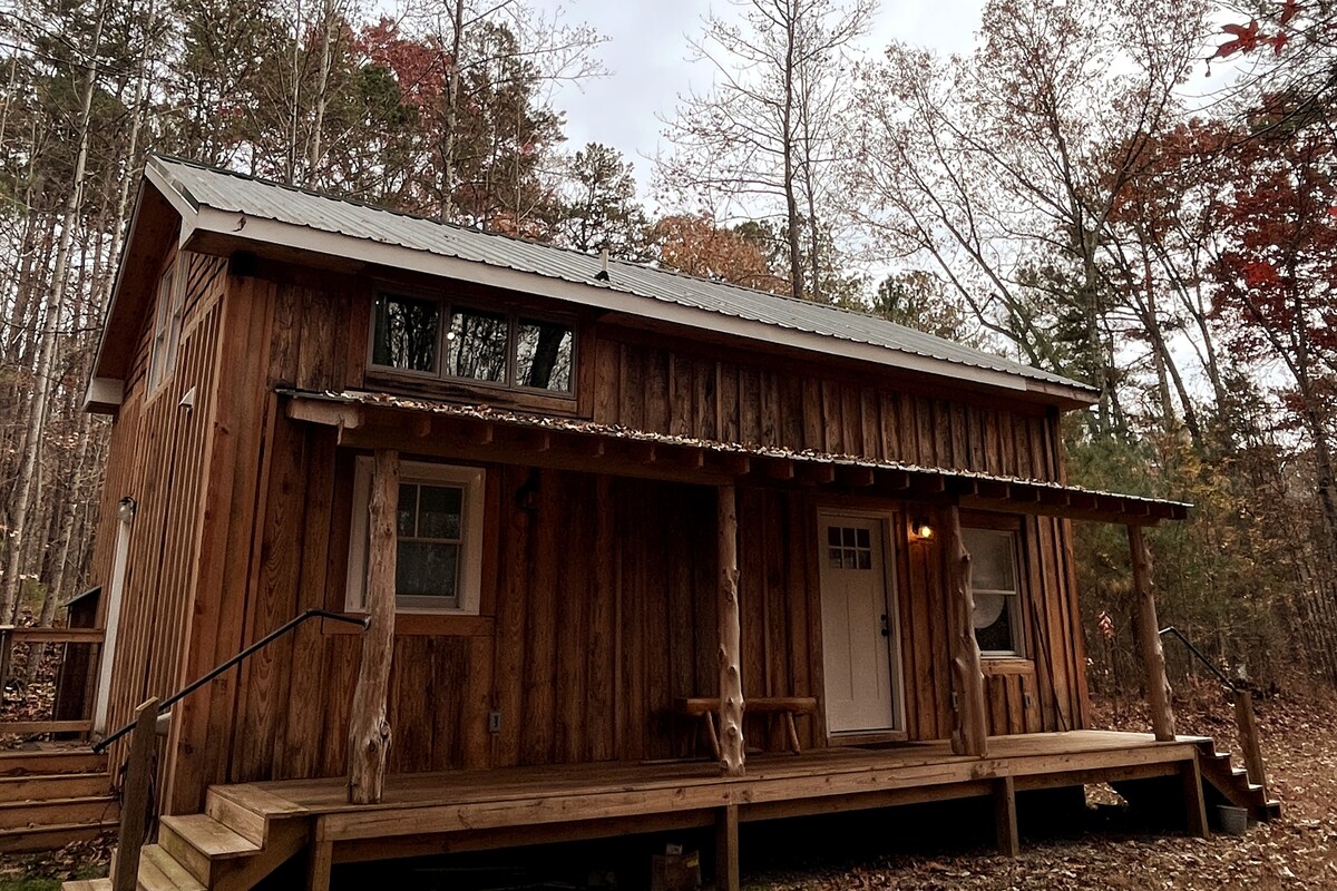 A rustic wooden cabin features a sloping metal roof and large front porch. Two windows allow natural light to enter, while steps lead up to the entrance. Surrounding trees reflect the colors of the season, enhancing the tranquil woodland setting.