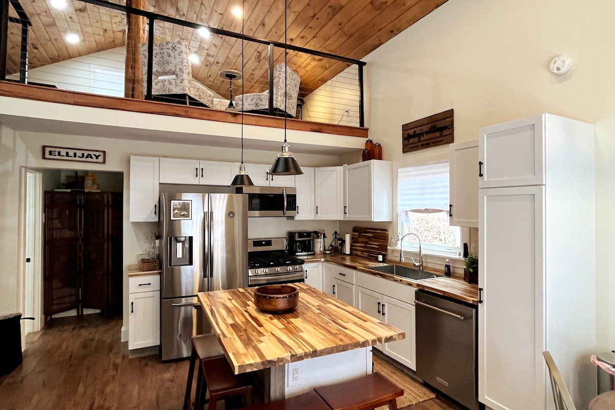 A modern kitchen space is showcased, featuring stainless steel appliances and a wooden island with high stools. Natural light fills the area, highlighting the white cabinetry and rustic wood accents, while a lofted seating area is visible above, outfitted with comfortable chairs.