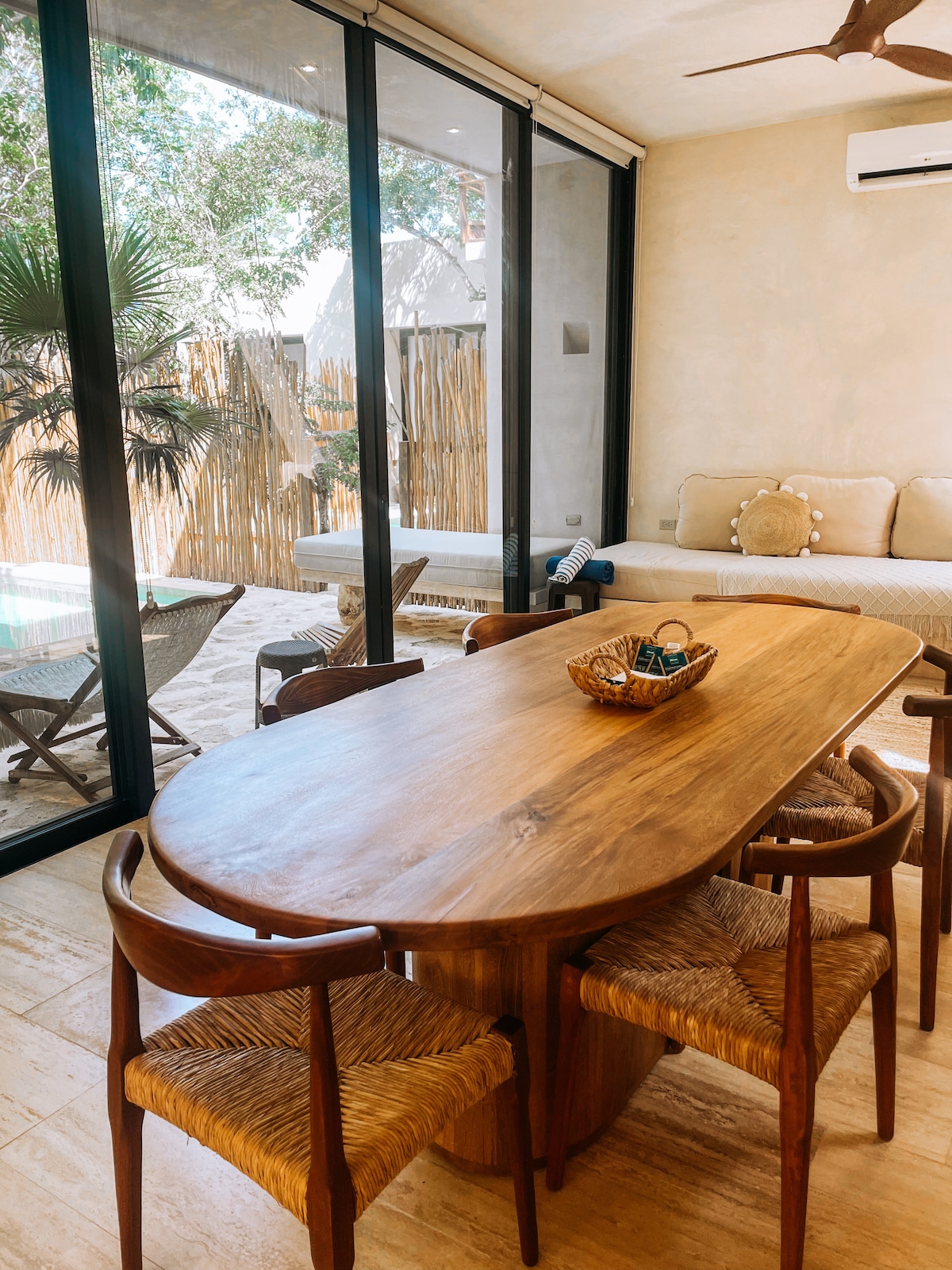A wooden dining table surrounded by four woven chairs is positioned in the living area. Floor-to-ceiling glass doors allow natural light to flow in, offering views of the outdoor space with a hammock and seating area.