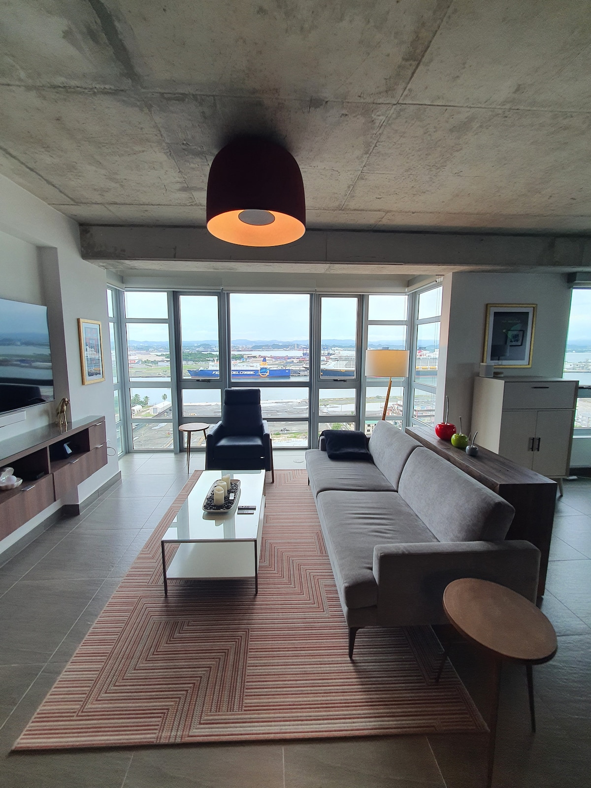 A contemporary living area features large windows that overlook the airport and cityscape. A gray sofa is positioned opposite a sleek coffee table, with a black armchair nearby. A floor lamp stands to the side, complementing a modern entertainment unit against the wall.