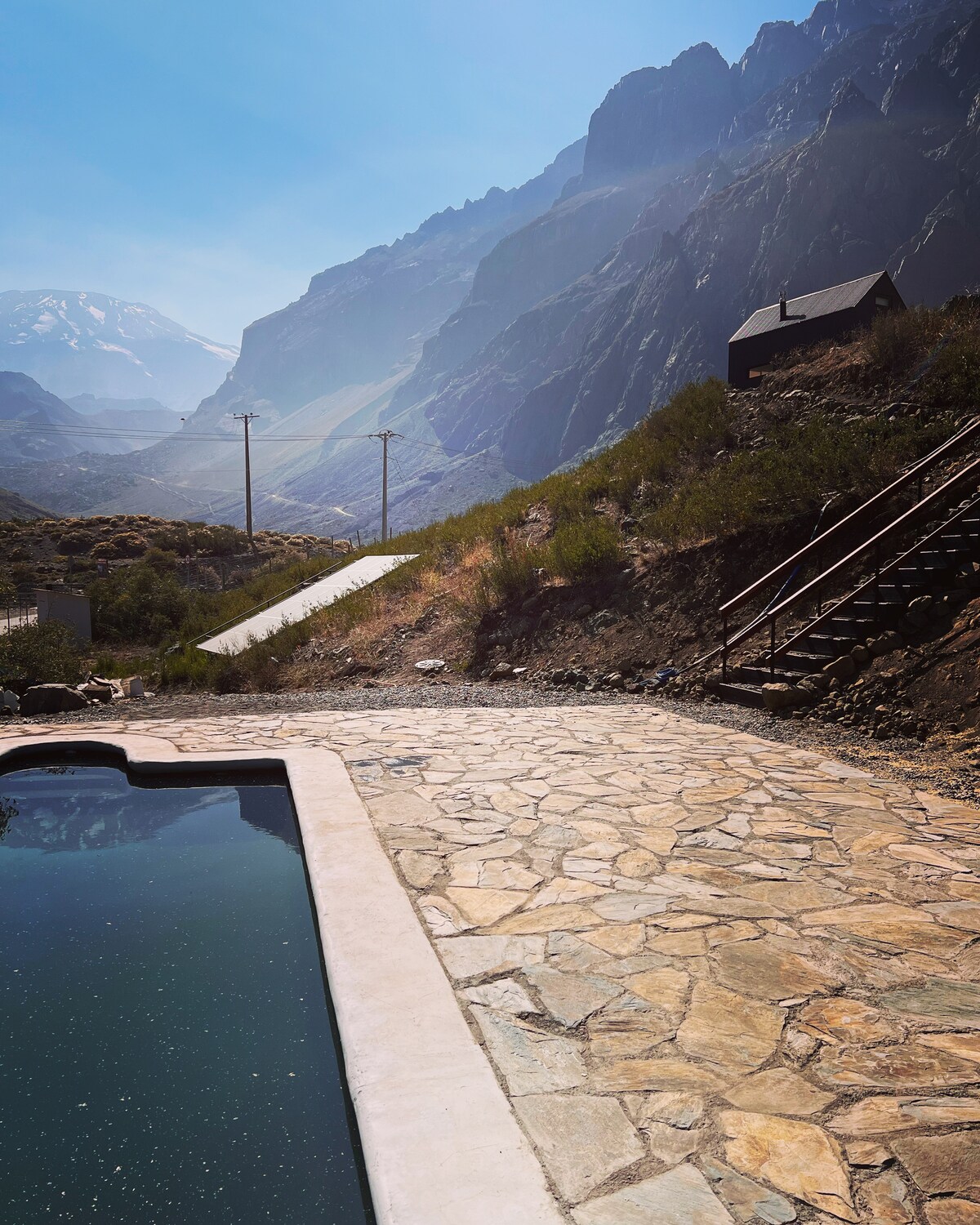A stone path leads to a tranquil pool, surrounded by rugged mountainous terrain. Wooden stairs ascend the hillside, connecting the area to a small cabin nestled among the rocks. Power lines stretch along the background, adding a hint of infrastructure to the natural landscape.
