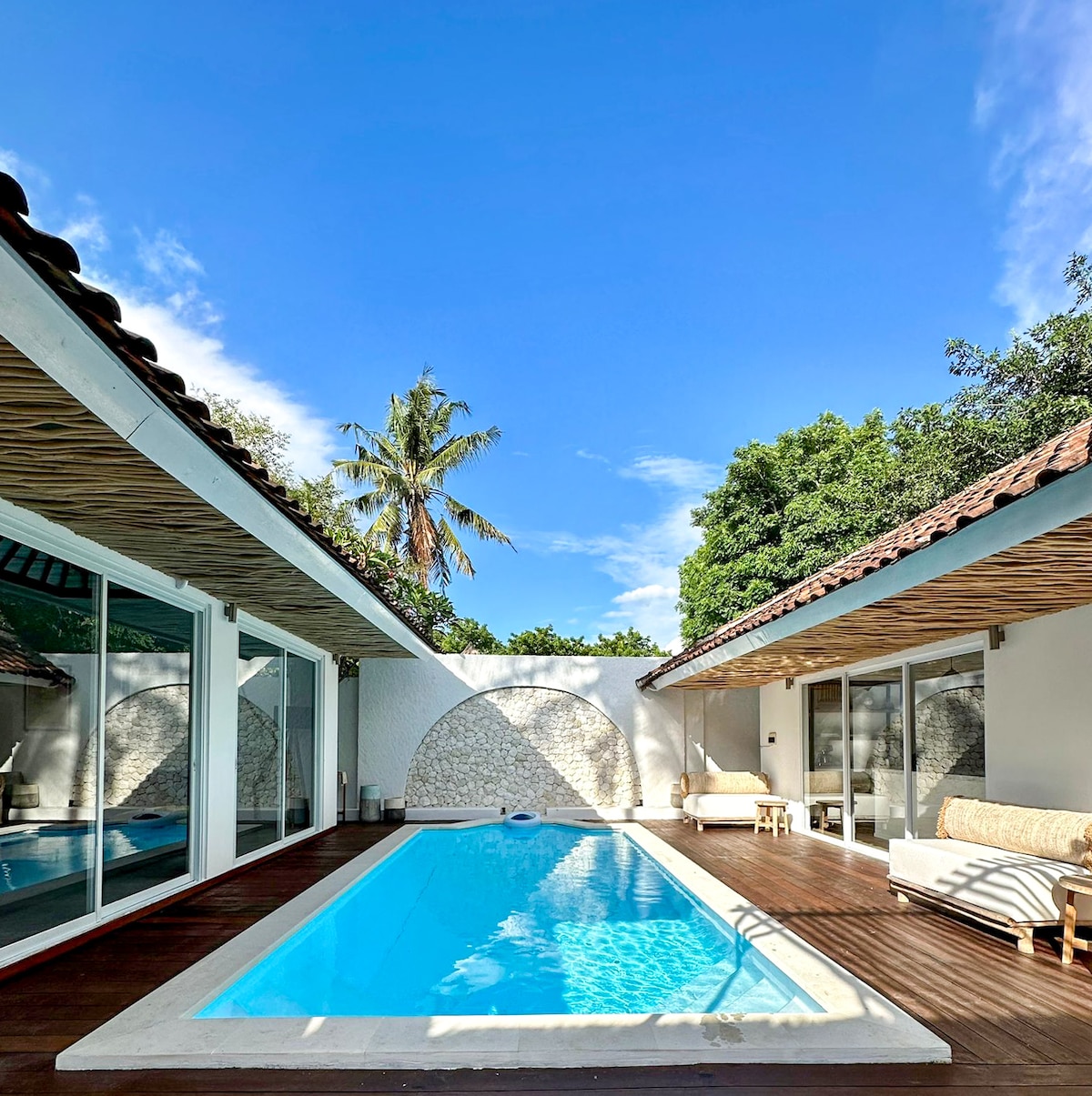 A serene swimming pool is set against a backdrop of lush greenery, flanked by open spaces showcasing modern design. Sunlight reflects off the water's surface, while comfortable seating is positioned nearby, creating an inviting area for relaxation.