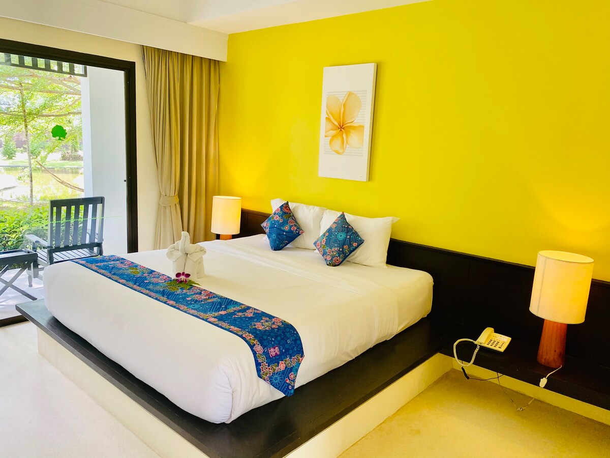 A spacious bed with a white coverlet is positioned against a bright yellow accent wall. Decorative pillows with floral patterns are featured. Soft lighting is provided by two bedside lamps, while a small table and a chair are visible in the corner, overlooking a garden view.