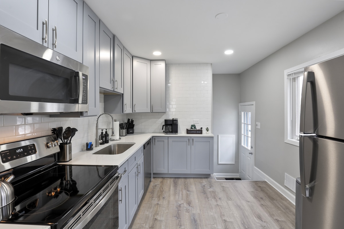 A modern kitchen is showcased, featuring light gray cabinetry and sleek stainless steel appliances. A white backsplash complements the countertop, while abundant natural light fills the space from nearby windows. An exit door leads to an outdoor area, enhancing convenience.