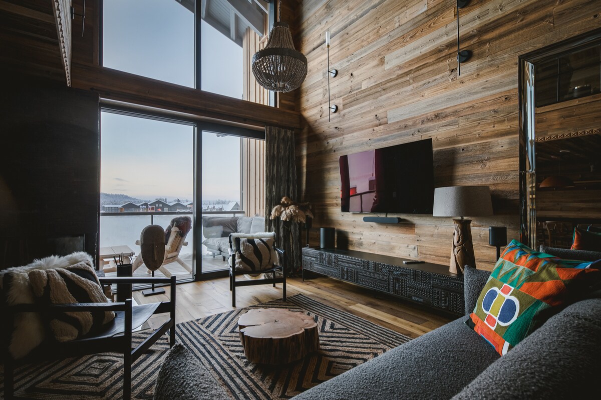 A spacious living area features a wooden accent wall and large windows offering views of the outdoors. A flat-screen television is mounted on the wall, accompanied by a stylish coffee table and comfortable seating arrangements, creating a relaxing atmosphere.