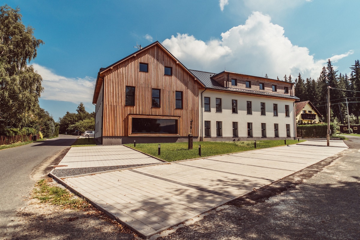 The exterior view of the building showcases a contemporary design featuring wooden paneling and a spacious façade. Surrounding greenery and a paved parking area create a welcoming entrance, complemented by clear blue skies and soft clouds.