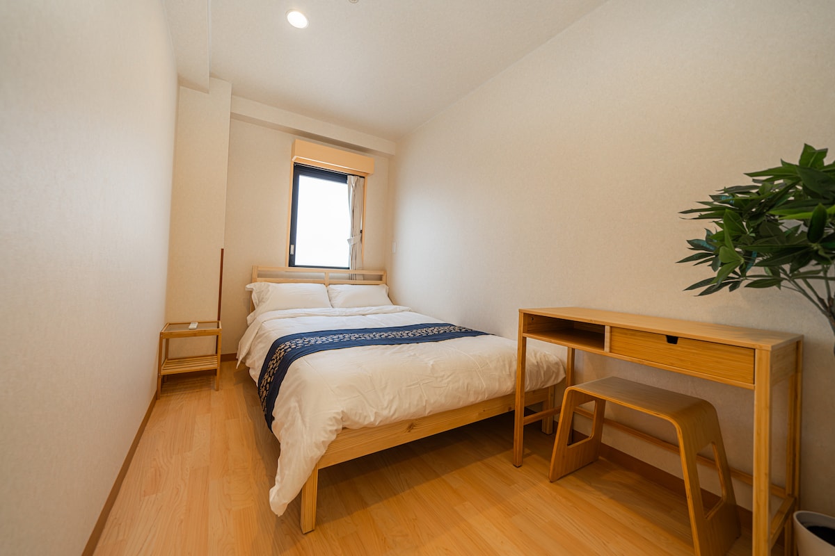 A bright bedroom features a spacious bed dressed in crisp white linens, accented by a dark blue throw. A small wooden desk and stool are positioned nearby, next to a window allowing natural light to illuminate the room. The light wood flooring adds a warm touch.