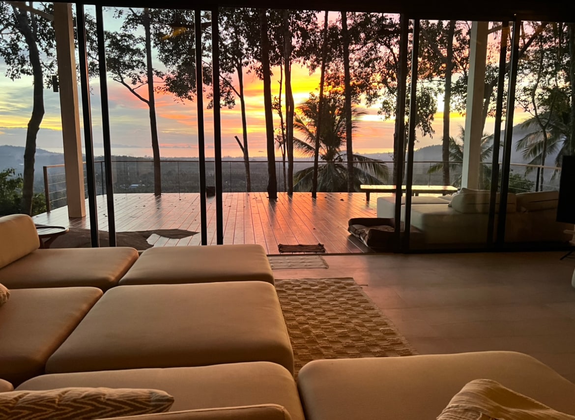 A spacious living area offers floor-to-ceiling glass walls, revealing a stunning view of a vibrant sunset surrounded by trees. Cozy seating is arranged invitingly, with a soft area rug adding texture to the contemporary decor and connecting indoor comfort with the outdoor scenery.