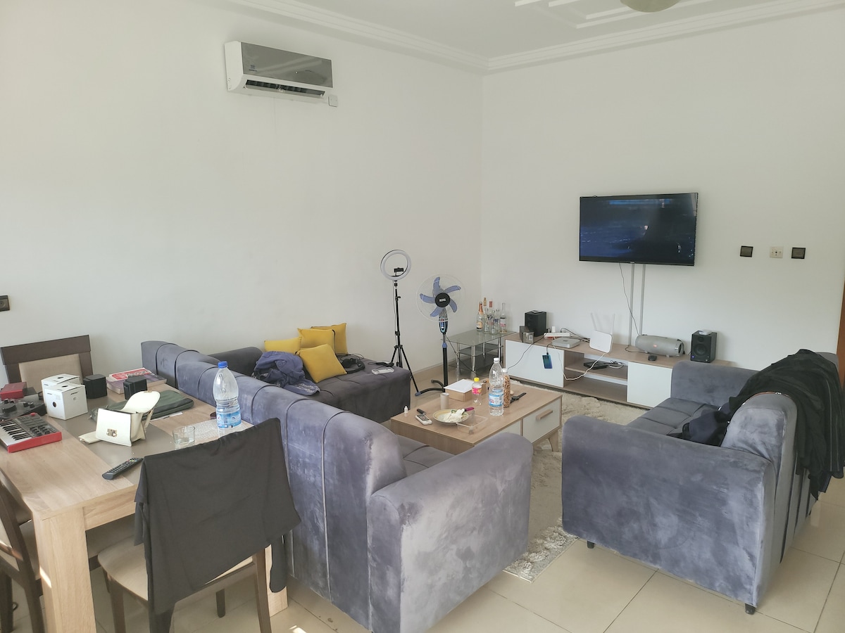A spacious living area features a plush sofa set and a wooden coffee table. A television is mounted on the wall opposite a decorative shelf. Various household items, including a fan and a water bottle, are visible, contributing to a relaxed atmosphere.