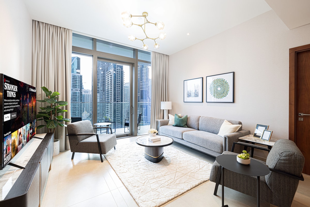 The spacious lounge area is furnished with a comfortable sofa and armchairs, complemented by a coffee table. Large windows provide a view of the city skyline. Decorative artwork adorns the walls, while indoor plants add a touch of greenery to the space.