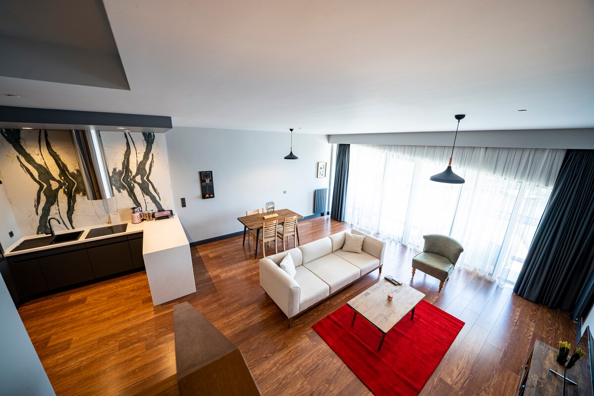 A spacious living area showcases a modern design with a light-colored sectional sofa and a wooden coffee table. Natural light filters through large windows, illuminating the dining area with a sleek table and chairs. A stylish green armchair adds a touch of comfort.