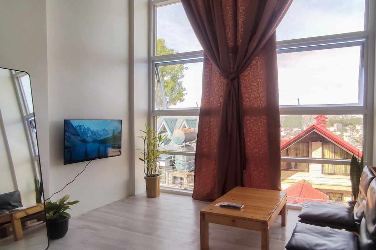A bright living area features large windows that offer a view of the city. A low wooden table is set on the light-colored floor, accompanied by a potted plant. A TV is mounted on the wall, and rich curtains frame the windows, creating a cozy ambiance.