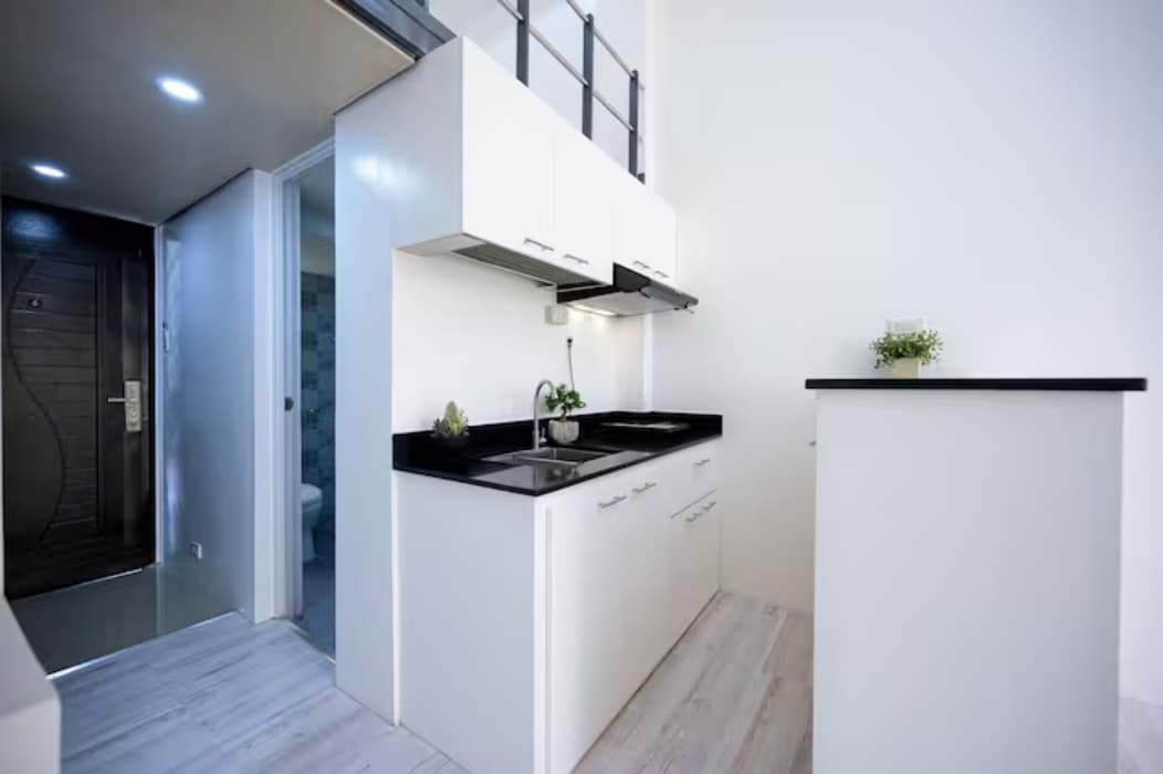 The kitchen area features minimalist cabinetry with sleek black countertops. Two burners are integrated beneath a pair of modern exhaust hoods. A small island can be seen, providing additional workspace. Space is well-lit by recessed ceiling lights, enhancing its clean and modern feel.