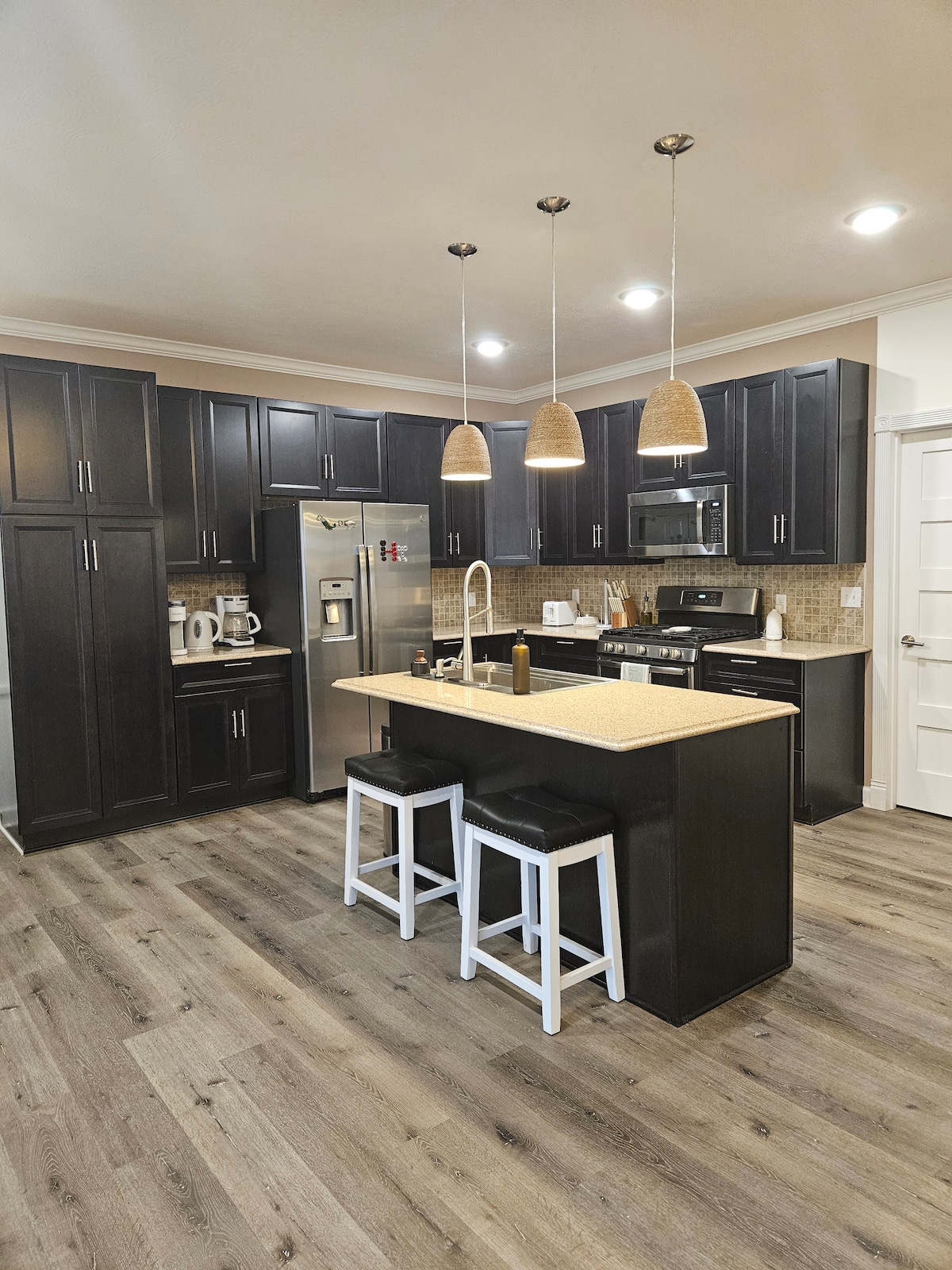 The kitchen features dark cabinetry and a central island with seating for two. Light fixtures hang above the island, and modern appliances, including a refrigerator, stove, and microwave, are arranged against the wall. A warm wood floor enhances the space.