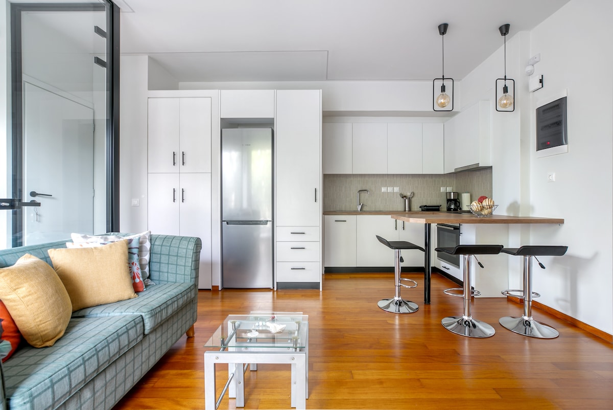 A modern open-plan living area is shown, featuring a blue-green sofa adorned with colorful cushions. A stylish dining table with high chairs sits adjacent to a contemporary kitchen equipped with stainless steel appliances, including a refrigerator. Natural light floods the space through large glass doors.