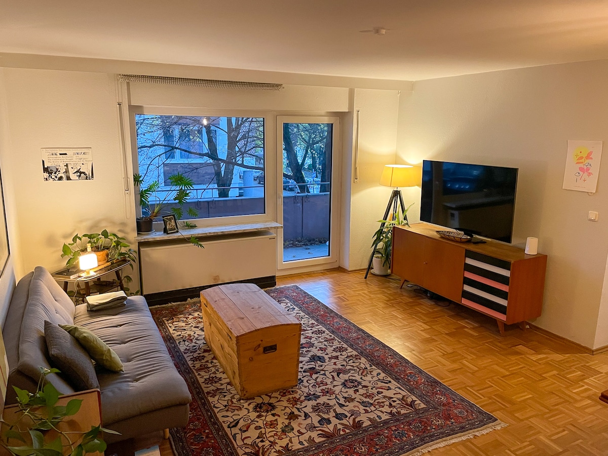 A spacious living area features a comfortable gray sofa and a wooden coffee table at its center. A large window allows natural light to fill the room, while a flat-screen television is mounted on a modern cabinet. Indoor plants add a touch of greenery.