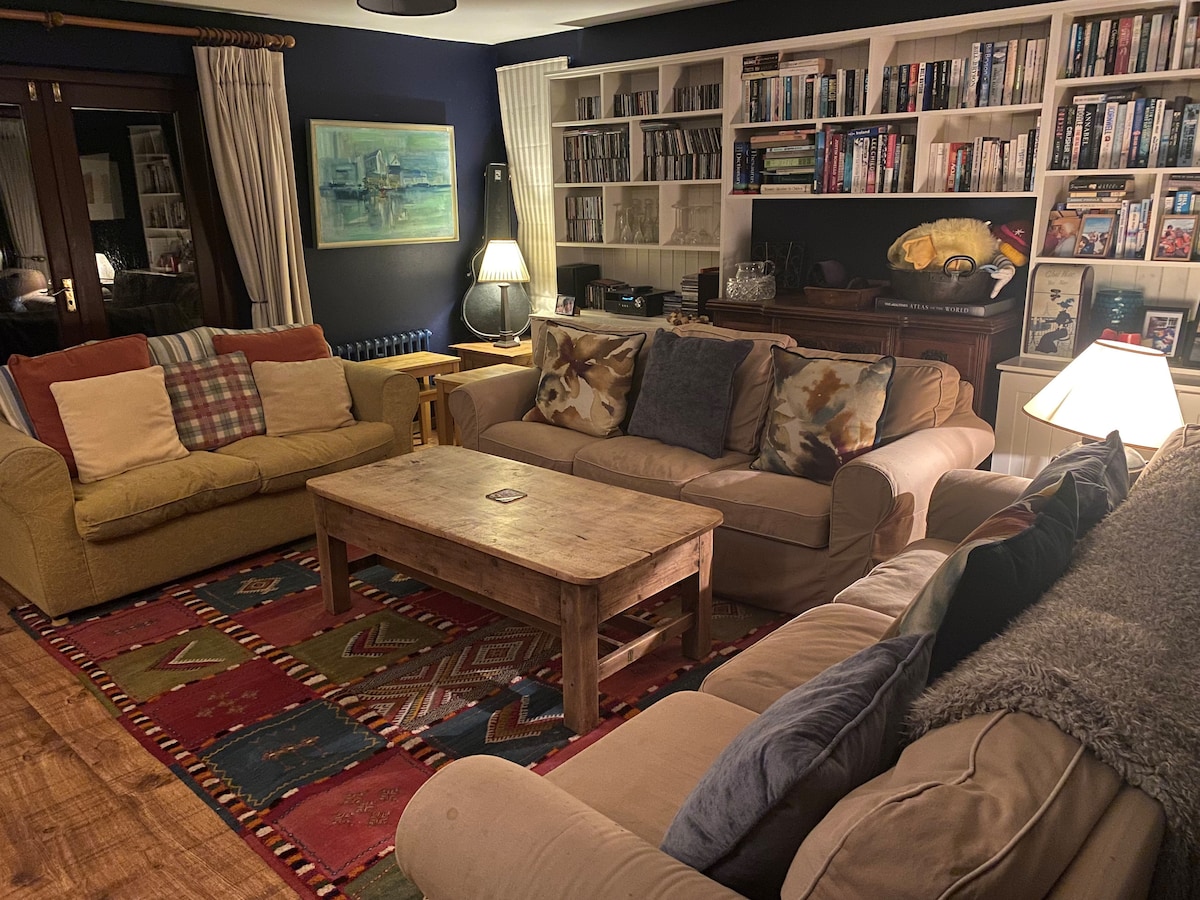 A spacious living room features two comfortable sofas in neutral tones, adorned with colorful decorative pillows. A wooden coffee table sits at the center, surrounded by a vibrant area rug. Shelves display books and DVDs, while soft lighting creates a cozy ambiance.