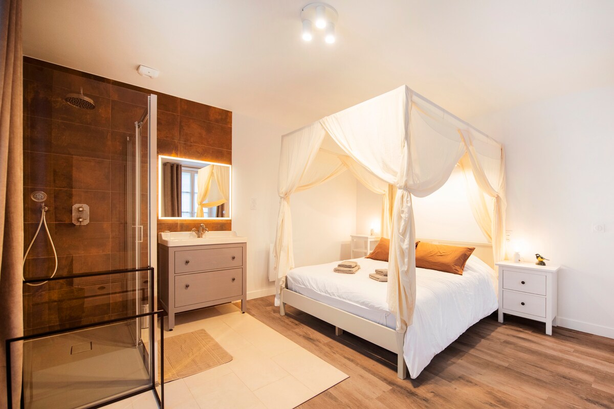 A spacious bedroom features a four-poster bed draped in light fabric, promoting a serene atmosphere. A modern shower area is visible, along with a sleek vanity. Warm lighting accents the decor, including a bedside table and a neutral-colored rug placed on the tiled floor.