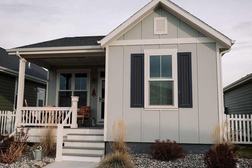 A light-colored cottage is shown from the front, featuring a small porch with two wooden chairs. A white railing and steps lead to the entrance. Decorative shutters frame the window, and low-maintenance landscaping with ornamental grasses adds visual interest.