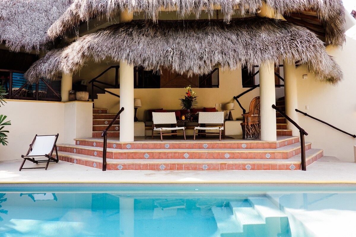 A spacious outdoor lounge area is positioned beneath a thatched roof, featuring comfortable seating arrangements. Steps lead down to a private swimming pool, reflecting the surrounding tropical landscape. Lush greenery frames the area, creating an inviting atmosphere for relaxation.