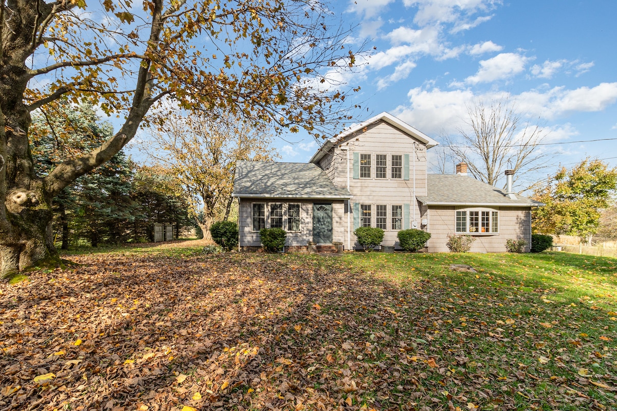 The farmhouse exterior is framed by autumn foliage, with leaves scattered across the green yard. A charming two-story design features large windows and a welcoming front entry. Surrounding trees and shrubs contribute to the rural aesthetic under a partly cloudy sky.