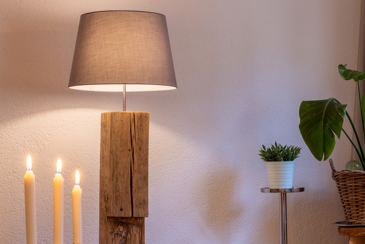 A wooden lamp with a gray shade stands on a textured base, providing soft illumination. Nearby, three white candles are positioned on a rustic surface, while a green plant in a white pot is placed next to a small table, enhancing the room's natural feel.