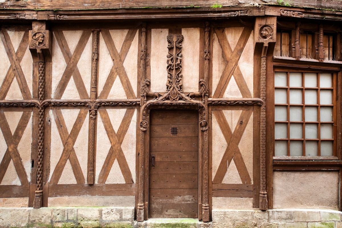 The image showcases a historic building façade featuring intricate wooden beams arranged in a diamond pattern. A wooden door with decorative carvings is situated at the center, surrounded by details that highlight craftsmanship. A small, barred window is visible to the right.