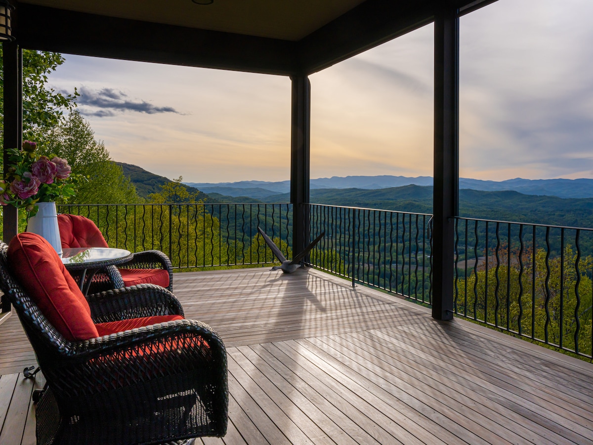 An outdoor porch features two wicker chairs with red cushions positioned on a wooden floor, offering expansive views of the surrounding mountains. A small table sits between the chairs, while the railing presents an unobstructed sight of the landscape under a softly lit sky.