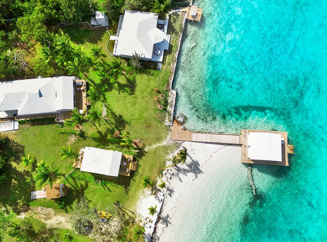 An aerial view showcases three cozy villas surrounded by lush greenery, with palm trees dotting the landscape. A wooden dock extends into the clear turquoise waters, leading to a beach that curves gently along the shoreline, highlighting the property’s proximity to the beach.