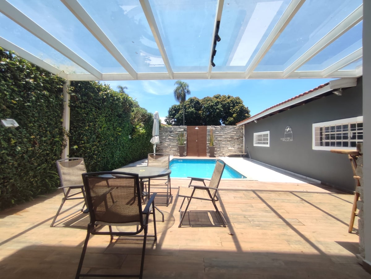 An outdoor patio area is presented with a clear roof, providing natural light. Four lounge chairs are arranged around a wooden deck, facing a swimming pool surrounded by greenery. A white umbrella stands beside the pool, enhancing the sunlit space.