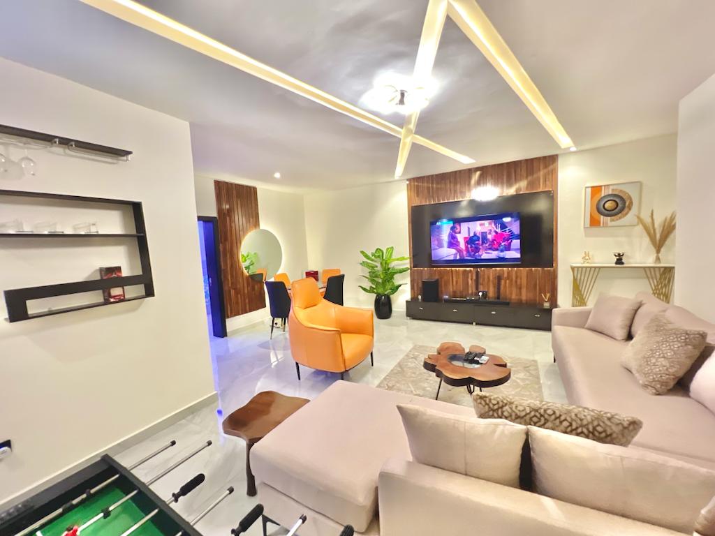 The living area features a spacious sofa paired with a unique coffee table. A vibrant orange chair adds a pop of color, while the entertainment unit displays a large television. Green plants provide a touch of nature, and stylish decor enhances the modern aesthetic.