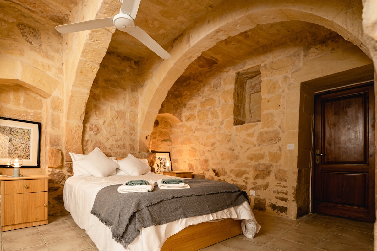 A cozy bedroom features stone walls with curved arches, creating a warm ambiance. A spacious bed is dressed in crisp linens, accompanied by neatly arranged towels and a softly draped blanket. A bedside table holds decorative items, while a door leads to the corridor.