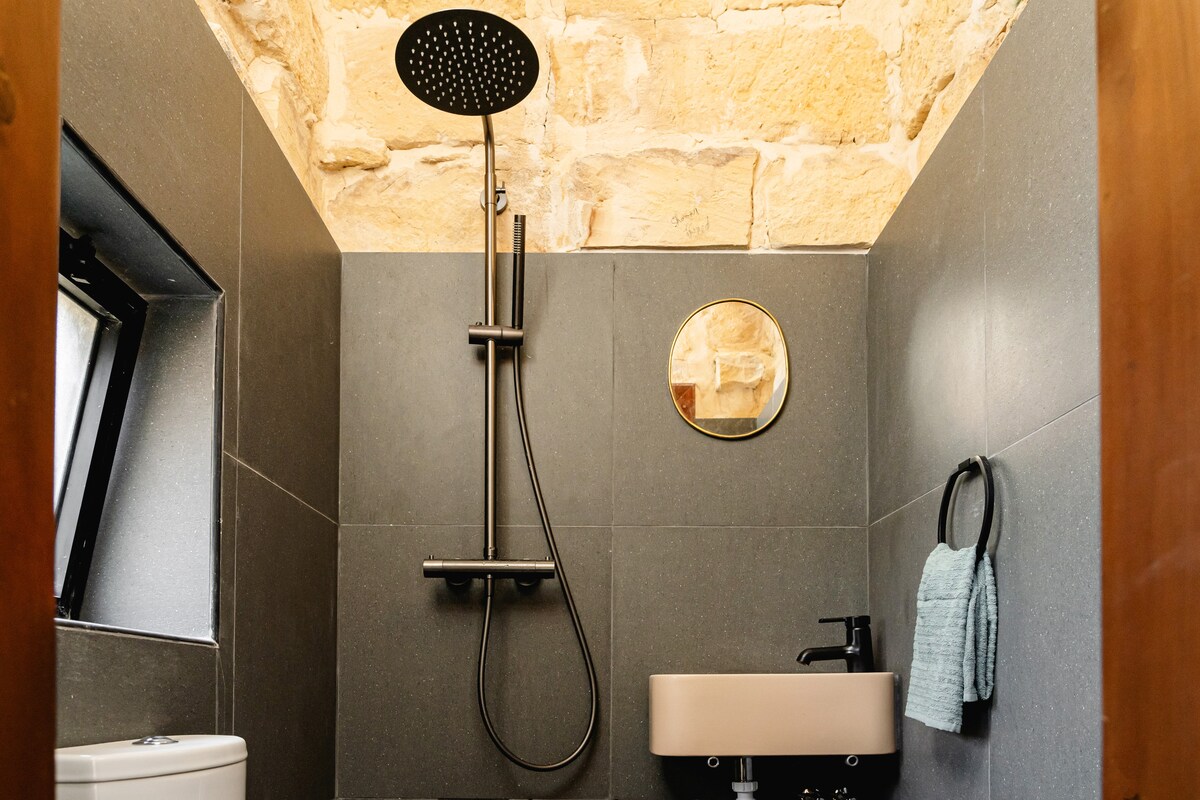 A modern bathroom features a rainfall showerhead mounted on a stone wall. A round mirror reflects soft light, complementing a sleek sink with a matte black faucet. A small towel hangs neatly by the wall, enhancing the room's functionality.