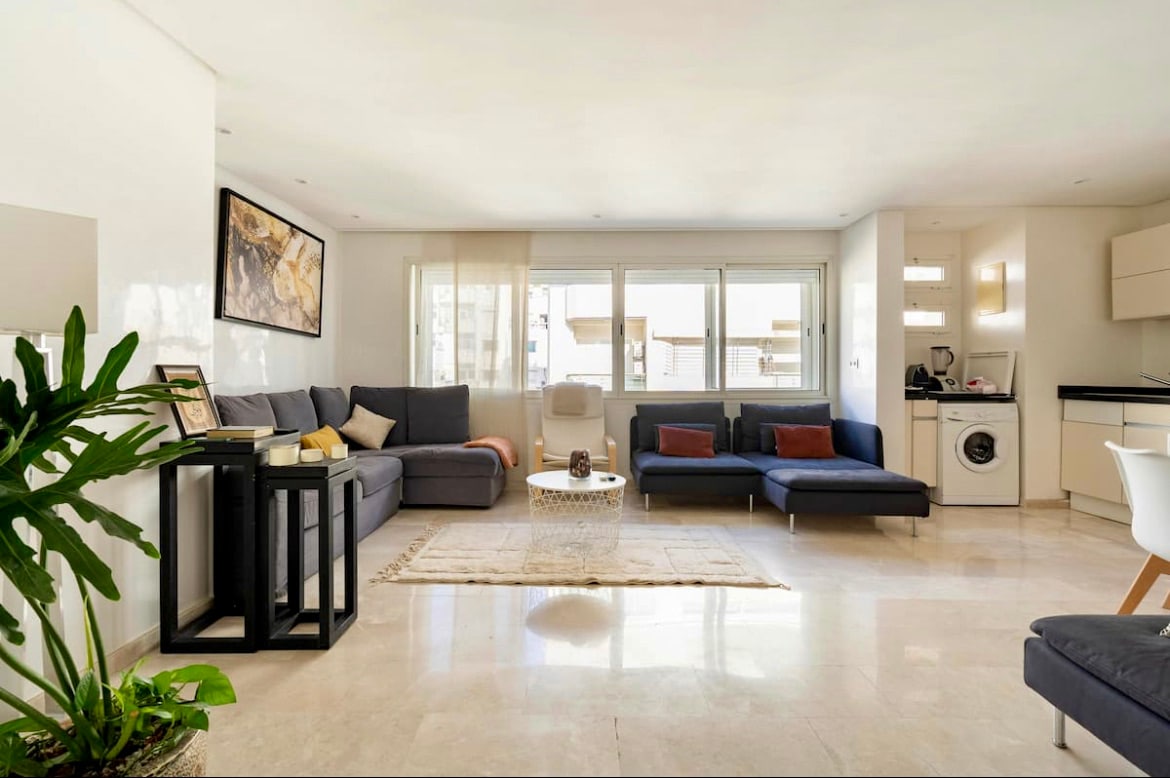 A spacious living area is composed of two comfortable sofas and a decorative coffee table. Natural light fills the space through large windows. A plant adds a touch of greenery, while a dining area and a kitchen are visible in the background.