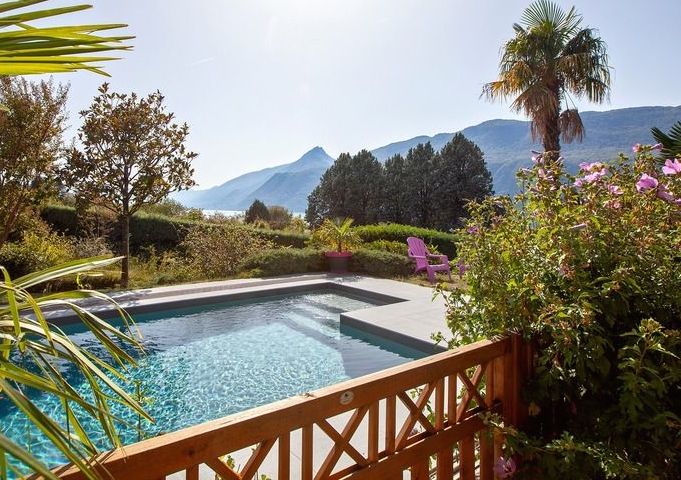 A serene outdoor scene features a clear swimming pool surrounded by lush greenery and colorful flowers. The distant mountains create a picturesque backdrop, while a single pink lounge chair invites relaxation. Sunlight illuminates the area, enhancing the tranquility of this inviting space.